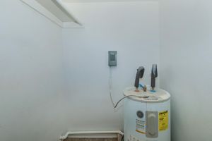 A small, unfurnished room with a white wall. In the corner, there is a water heater with visible pipes and valves. On the adjacent wall, there is a small electrical panel. The floor is covered with a simple carpet, and the overall ambiance is plain and utilitarian.