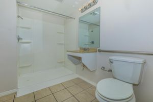 A clean, modern bathroom featuring a walk-in shower with a clear glass door, a small sink with a countertop, a large mirror, and a white toilet. The floor is tiled. The walls are painted in a light color, enhancing the brightness of the space.