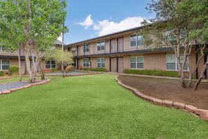 A well-maintained courtyard featuring green grass and landscaping with shrubs. The area has two-story apartment buildings with balconies, surrounded by pathways and decorative stone borders. The sky is partly cloudy, creating a pleasant atmosphere for residents.