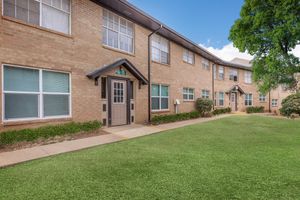 A well-maintained, two-story brick apartment building with multiple windows and a central entrance. The exterior features neatly trimmed grass and small shrubs, creating a welcoming atmosphere. The sky is clear with a few clouds, adding to the bright appearance of the property.