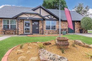 A single-story brick building with a slate roof, featuring a front entrance marked "1332" and two benches on either side. In front, there's a landscaped area with decorative rocks, plants, and a small pot. A red flag stands to the side, and the setting includes greenery and a blue sky.