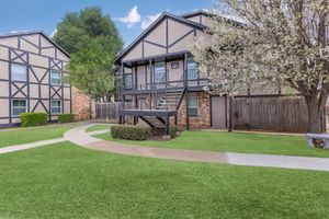 Two multi-story residential buildings with a modern architectural style, surrounded by well-maintained green lawns and flowering trees. A pathway leads toward the entrance of the building on the right, which features a staircase and balcony. The scene is set under a clear blue sky.