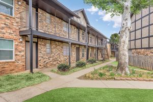 A view of a brick apartment community with two stories, featuring balconies, a landscaped pathway, and green grass. A large tree is visible in the foreground, alongside flower beds and shrubbery, creating a welcoming outdoor space. The sky is partly cloudy.
