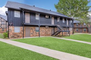 A two-story apartment building with black wooden siding and brick accents. It features multiple doors, windows, and a staircase. The building is surrounded by well-maintained green grass and a clear blue sky in the background.