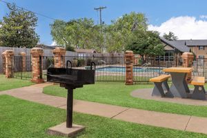 A well-maintained outdoor area featuring a black charcoal grill in the foreground, a wooden picnic table, and a fenced-in swimming pool surrounded by greenery and trees. The sky is clear, and the overall space appears inviting for gatherings and relaxation.