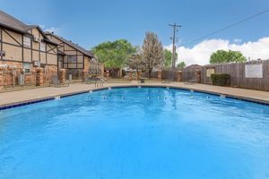 A clear blue swimming pool surrounded by a concrete deck. The area features several lounge chairs and a landscaped outdoor space with trees and a fence in the background. The sky is bright with a few clouds, creating a pleasant atmosphere for relaxation.