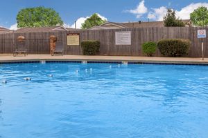 A clear blue swimming pool surrounded by a wooden fence. In the background, there are several trees and houses. White buoys are visible in the water, and there are two lounge chairs on the poolside. Signs indicate pool rules, including a "No Diving" warning.