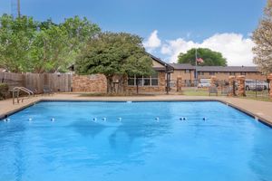A clear blue swimming pool surrounded by a patio area with lounge chairs. Lush green trees and a brick building are visible in the background, along with a flagpole displaying the American flag. The scene is bright and inviting, ideal for relaxation and recreation.