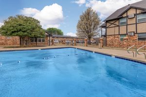 A clear blue swimming pool surrounded by grass and trees, with brick buildings in the background. The sky is partly cloudy, creating a peaceful outdoor setting. There are lounge chairs near the pool and a few people visible in the distance.