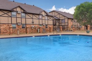 Swimming pool with clear blue water in front of two-story apartment buildings featuring brick and stucco exteriors. The area is surrounded by a fence, with lounge chairs and a shaded picnic table nearby, under a blue sky with a few clouds.