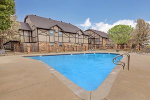 A clear blue swimming pool surrounded by a concrete patio, with lounge chairs scattered around. In the background, there are two-story apartment buildings featuring a mix of brick and wood siding, and trees providing some greenery. The sky is partly cloudy, adding a bright ambiance to the scene.