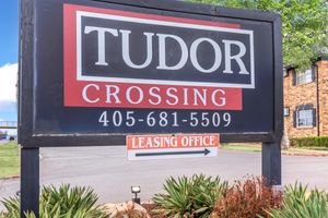 Sign for Tudor Crossing, displaying the name prominently with contact number 405-681-5509. An arrow indicates the direction to the leasing office. The background features well-maintained greenery and a clear blue sky.