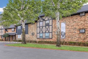 A brick building with a mix of stone and timber accents, featuring large windows and air conditioning units. The front is landscaped with grass and trimmed hedges. Visible trees line the pathway, and a sign is mounted on the façade, indicating the property name or purpose.
