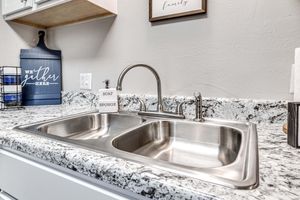 A modern kitchen sink featuring a double basin with a polished stainless steel finish. The countertop is made of light granite with dark speckles. There is a decorative soap dispenser labeled "SOAP" and a sponge holder. A blue wall sign in the background reads "we gather here."