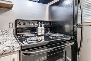 A clean black kitchen stove with four burners on a granite countertop, accompanied by a small container of bottles in the center. To the right is a stainless steel refrigerator, creating a modern kitchen environment. The wall is painted white, contributing to the bright, tidy space.