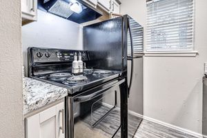 A modern kitchen featuring a black stove and oven with a granite countertop, a black refrigerator, and overhead cabinets. The kitchen has a window with blinds allowing natural light, and the walls are painted a light color. The space appears tidy and well-organized.
