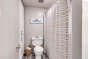 A cozy bathroom featuring a white toilet, a shower curtain with a striped design, and a woven basket. The walls are painted in a soft color, and a decorative artwork depicting a sea creature adds a touch of charm. The flooring is wood laminate, contributing to a warm atmosphere.