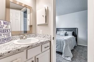 A view of a bathroom with a modern sink and granite countertop, featuring a large mirror above the sink. In the background, a bedroom is visible with a neatly made bed and light-colored walls. A decorative sign next to the sink reads "Do Not Feed the Sharks or Tease the Mermaids."