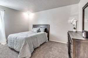A softly lit bedroom featuring a cozy bed with light blue bedding, a wooden headboard, and accent pillows. There's a dresser against the wall, a decorative plant, and a window with sheer curtains allowing natural light to filter in, creating a calm and inviting atmosphere.