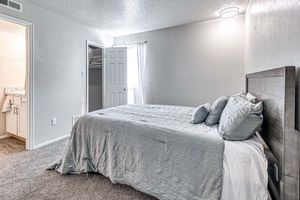 A cozy bedroom featuring a neatly made bed with grey bedding and decorative pillows. There is a door leading to a closet on the left, and a window with sheer curtains allowing natural light to enter. The walls are painted light grey, and the carpeted floor adds warmth to the space.