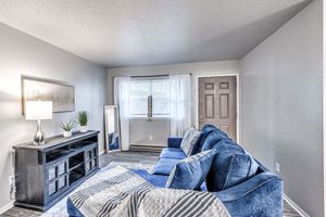 A cozy living room featuring a blue couch, a decorative throw blanket, and a small side table with a lamp. A large mirror stands beside the couch, while a potted plant adds a touch of greenery. Natural light streams in through a window with sheer curtains, and a door is visible in the background. Earth-toned flooring completes the space.