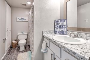 A bright and clean bathroom featuring a white vanity with a granite countertop, a mirror above, and a decorative sign that reads "Do Not Feed the Sharks or Tease the Mermaids." A toilet is visible beside a shower with a striped curtain, and a woven basket is on the floor, adding a cozy touch to the space.
