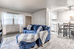Cozy living area featuring a blue sectional sofa with a throw blanket, a decorative rug, and a window with sheer curtains. In the background, a kitchen space with white cabinetry and a dining table set for four is visible, creating an inviting atmosphere.