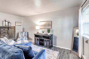 A cozy living room featuring a blue sofa and chair, a wooden cabinet, a lamp on a table, and decorative plants. A mirror reflects part of the space, and natural light filters through a window with white curtains, creating a warm and inviting atmosphere.