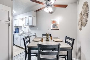 A cozy dining area featuring a wooden table set for four, with plates and candles. A ceiling fan hangs above. In the background, a modern kitchen with white cabinets, a marble countertop, and artwork on the wall. The space is well-lit and has a warm, inviting atmosphere.