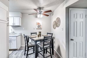 A dining area featuring a wooden table with four chairs, a ceiling fan, and a light fixture. The space has light-colored walls and a large window. On the table are placemats, and a decorative item is placed in the center. A framed artwork is hung on the wall, adding a touch of decor.