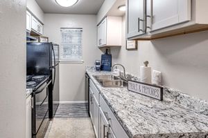A compact kitchen featuring white cabinets and a dark black stove. The countertop is made of granite with a silver sink. A window allows natural light, and a decorative sign reads "Meals & Memories." The floor has a light-colored tile, creating a cozy and inviting atmosphere.