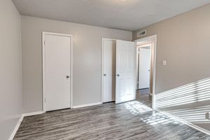 A well-lit room featuring light gray walls and laminate flooring. There are two closed doors and one partially open door leading to another space. Sunlight casts shadows across the floor from the door frame.