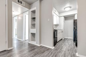 A view of a modern kitchen with white cabinets and a black refrigerator, adjacent to a hallway leading to other rooms. The flooring is light-colored and has a wood-like appearance. The walls are painted a neutral shade, contributing to a bright and open feel.