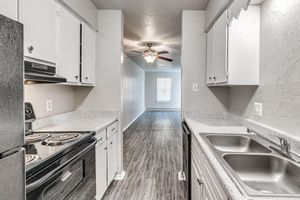 A modern kitchen featuring black appliances, white cabinets, and a double sink, with a view of a living area in the background. The flooring is a light wood laminate, and there is a ceiling fan in the living space. Natural light comes through a window at the far end.