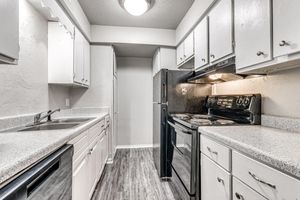 A modern kitchen featuring white cabinets, a black refrigerator, and an oven with a smooth top. The countertops are speckled gray, and there is a single basin sink. The floor has a wood-like appearance, and the room is well-lit with overhead lighting.