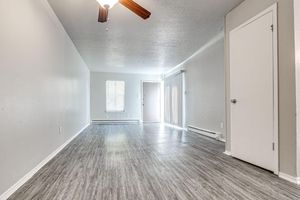 A spacious, minimalist interior of a room featuring light gray walls, a ceiling fan, and laminate flooring. There are windows allowing natural light, and a white door leads to another area.