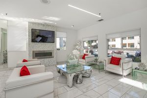 A modern, stylish waiting area featuring two white armchairs with red cushions, a glass coffee table with a decorative vase, and a flat-screen TV mounted on a textured wall. Large windows allow natural light and offer a view of the street outside. Bright, minimalist decor with sleek lines and a clean aesthetic.