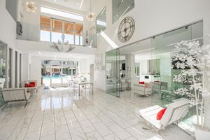 Bright and spacious modern interior featuring high ceilings, large glass doors, and sleek white decor. The open floor plan includes a glass dining table, minimalist furniture in white and red accents, and contemporary lighting fixtures. A pool can be seen through the large windows, enhancing the airy atmosphere.
