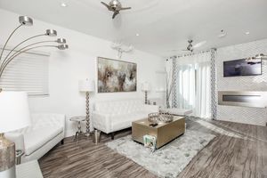 Bright and modern living room featuring a white leather sofa and chairs, a stylish coffee table, and contemporary decor. The space includes a large artwork on the wall, decorative lamps, a TV mounted above a sleek fireplace, and large windows with sheer curtains allowing natural light to fill the room.