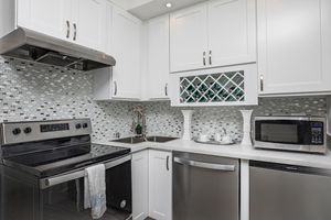 A modern kitchen featuring white cabinetry, a sleek stainless steel stove, microwave, and dishwasher. The backsplash is made of small mosaic tiles in gray and white tones. A small sink is located next to the stove, with a decorative wine rack above, displaying bottles of wine. A dish towel hangs from the stove.