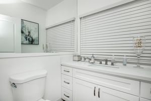 A clean and modern bathroom featuring a white toilet, a large sink with a mirror above, and cabinetry. Light-colored walls and blinds allow natural light, enhancing the spacious feel. A decorative glass with a message adds a personal touch to the sink area.