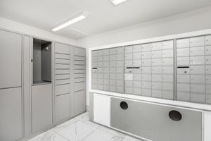 A modern mailroom with gray metal mailboxes, featuring a bank of small compartments for secure mail delivery. There is a larger open compartment on the left side and illuminated by overhead lights, with a clean, white wall and marble floor creating a bright and organized atmosphere.