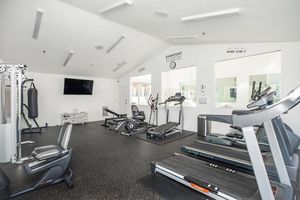 A bright and modern gym interior featuring various exercise equipment, including treadmills, stationary bikes, and strength machines. The space has large mirrors and a television on the wall, with motivational quotes displayed. The flooring is dark and rubberized, providing a clean and inviting workout environment.