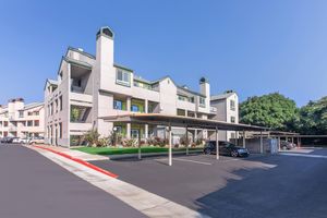 A modern apartment community featuring a light-colored building with multiple levels, balconies, and green accents. Covered parking is visible in the foreground, along with an asphalt driveway. Lush greenery surrounds the premises under a clear blue sky.