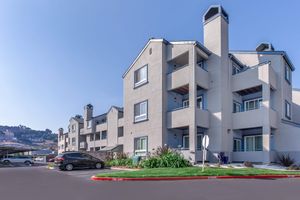A modern, multi-story residential building with a light gray exterior, featuring balconies and angled roofs. The scene includes parked cars, landscaped greenery, and a clear blue sky in the background, with hills visible at a distance.