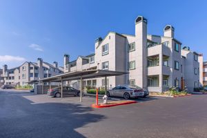 A modern residential apartment community featuring multiple beige buildings with triangular roofs and balconies. In the foreground, there are covered parking spaces and parked cars, along with landscaped areas with flowers and foliage under a clear blue sky.