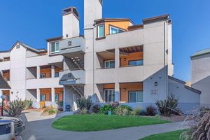 A modern apartment building with three floors, featuring a light gray exterior and orange accents. It has multiple balconies, situated around a landscaped area with grass and decorative plants. A pathway leads through the greenery, and a few vehicles are parked nearby. Bright blue sky overhead.