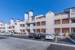 A modern apartment community featuring three stories with balconies, painted in light colors with orange accents. The building is surrounded by parking spaces, with several cars parked in front. The area includes desert landscaping with shrubs and succulents under a clear blue sky.