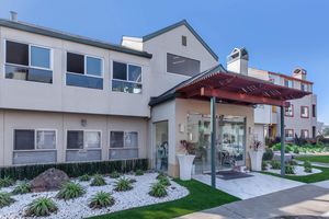 Modern building exterior featuring a glass entrance canopy supported by wooden beams. The facade has large windows and a mixture of light-colored walls, with neatly landscaped plants and decorative rocks in the foreground. Clear blue sky serves as a backdrop.