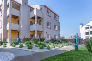 A modern multi-story apartment building with colorful accents, surrounded by landscaped greenery and decorative rocks. A pathway leads to the entrance, with a blue decorative feature in the foreground and a grassy area nearby. Clear blue skies are visible overhead.